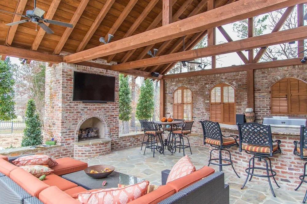 Outdoor covered patio with brick walls, wooden roof structure, ceiling fans, a flat-screen TV, a brick fireplace, a seating area with cushioned wicker furniture, a dining table with chairs, windows with shutters, and an outdoor kitchen area with a grill.