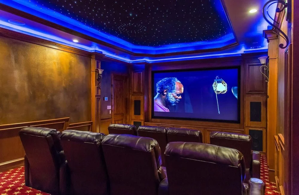 Home theater room with leather recliners, wooden panel walls, a large screen showing a man and an lantern, starry ceiling, and blue LED lighting.
