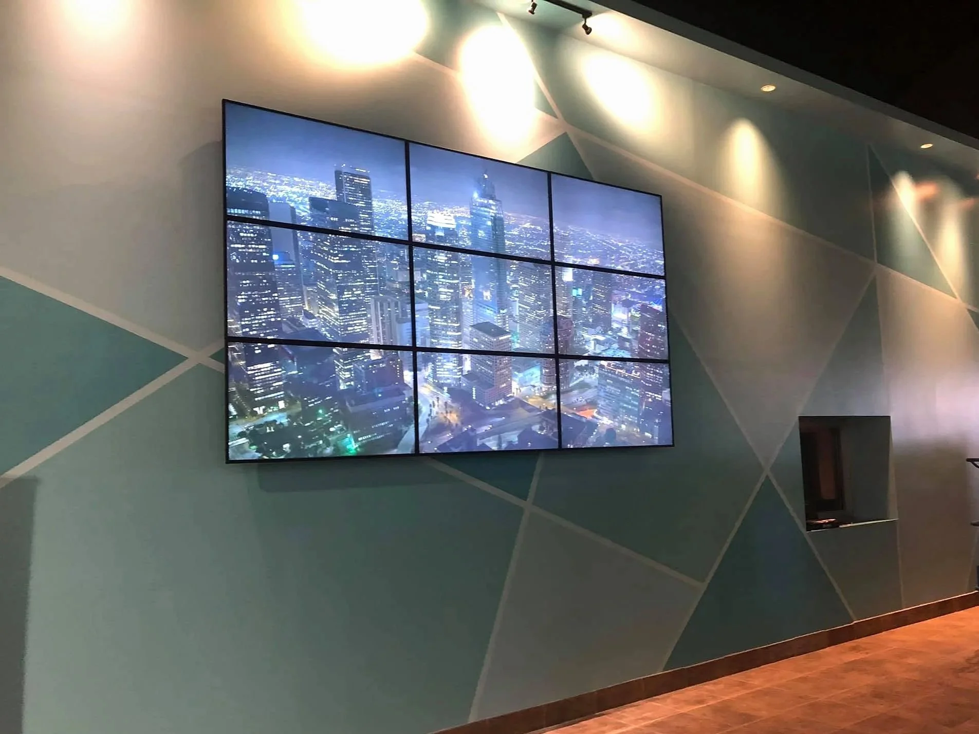 Interior of a modern building with a large digital display showing a nighttime cityscape of tall skyscrapers and city lights. The wall has a geometric pattern painted in shades of teal and gray. There are spotlights on the ceiling.