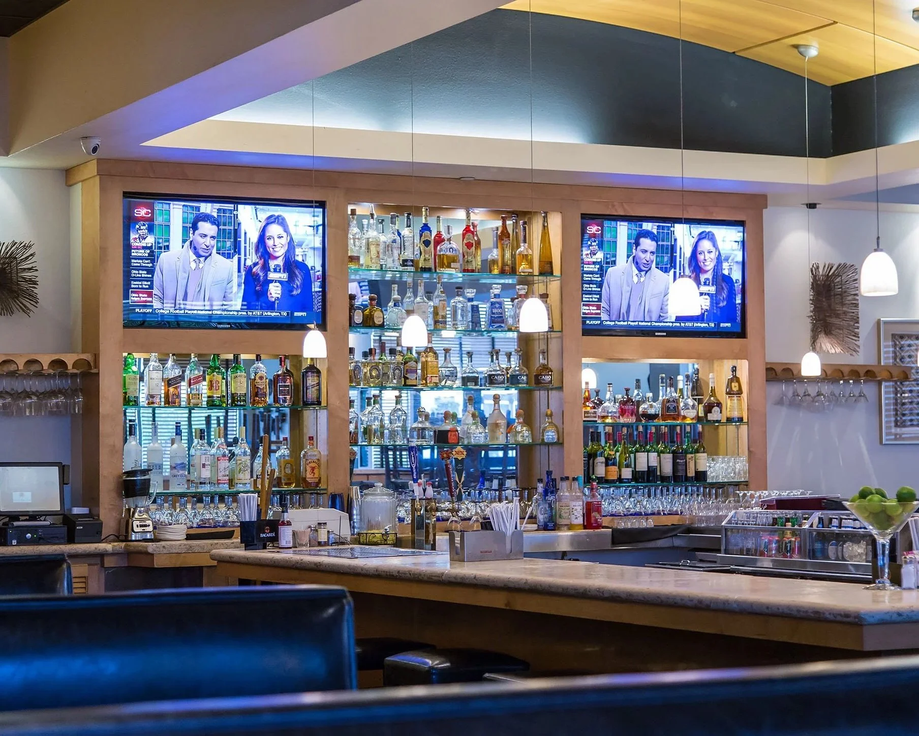 A bar with bottles of liquor on shelves, two TV screens showing news anchors, and pendant lights hanging from the ceiling.