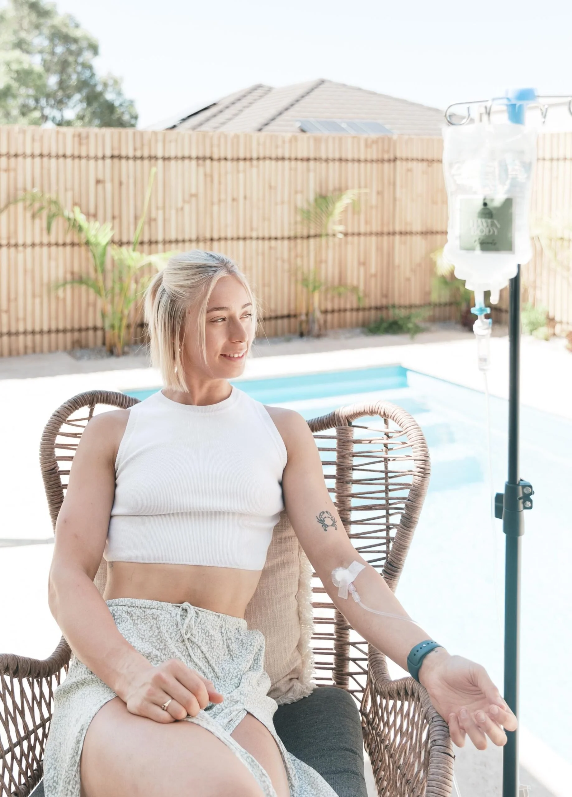 A woman sits in a wicker chair by a pool, receiving an IV drip, smiling, with a tattoo on her upper arm, wearing a white crop top and patterned shorts, outside in a backyard with a wooden fence and house in the background.