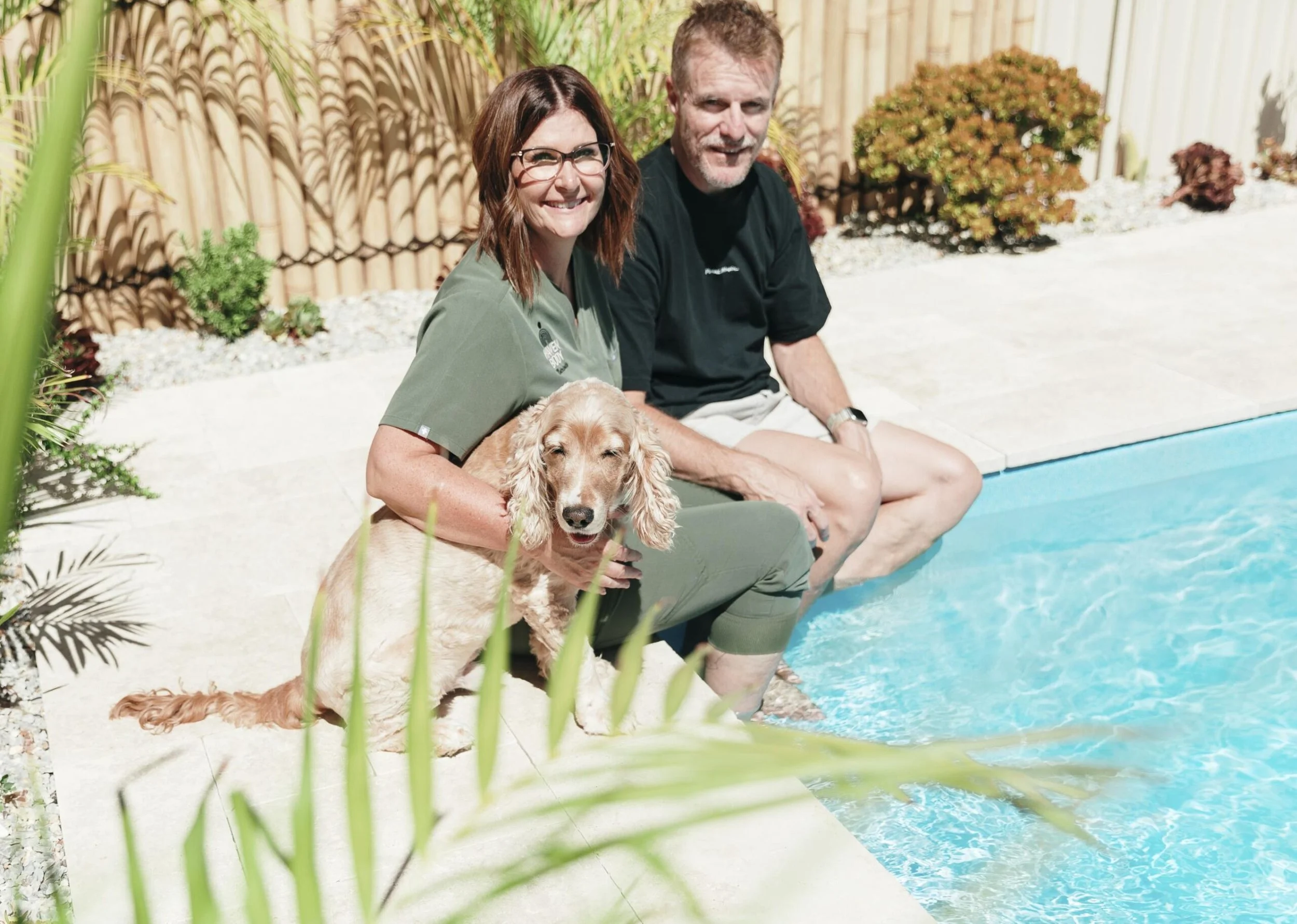 Smiling woman and man sitting by a pool with a dog on her lap, surrounded by plants and a bamboo fence.