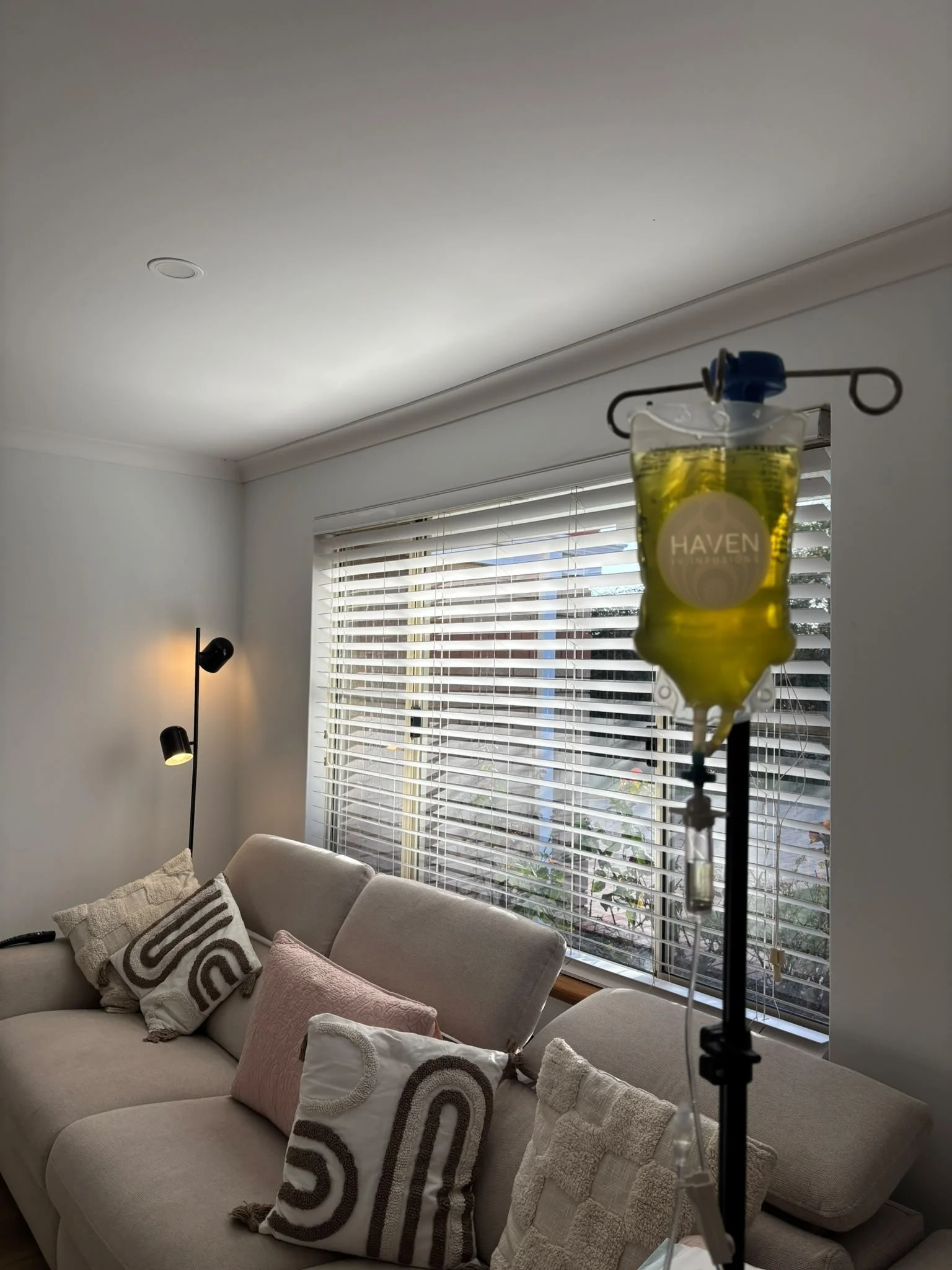 Living room with a beige sofa decorated with patterned and textured throw pillows, a side lamp, a wall with white blinds covering a window, and a medical IV drip stand in the foreground.