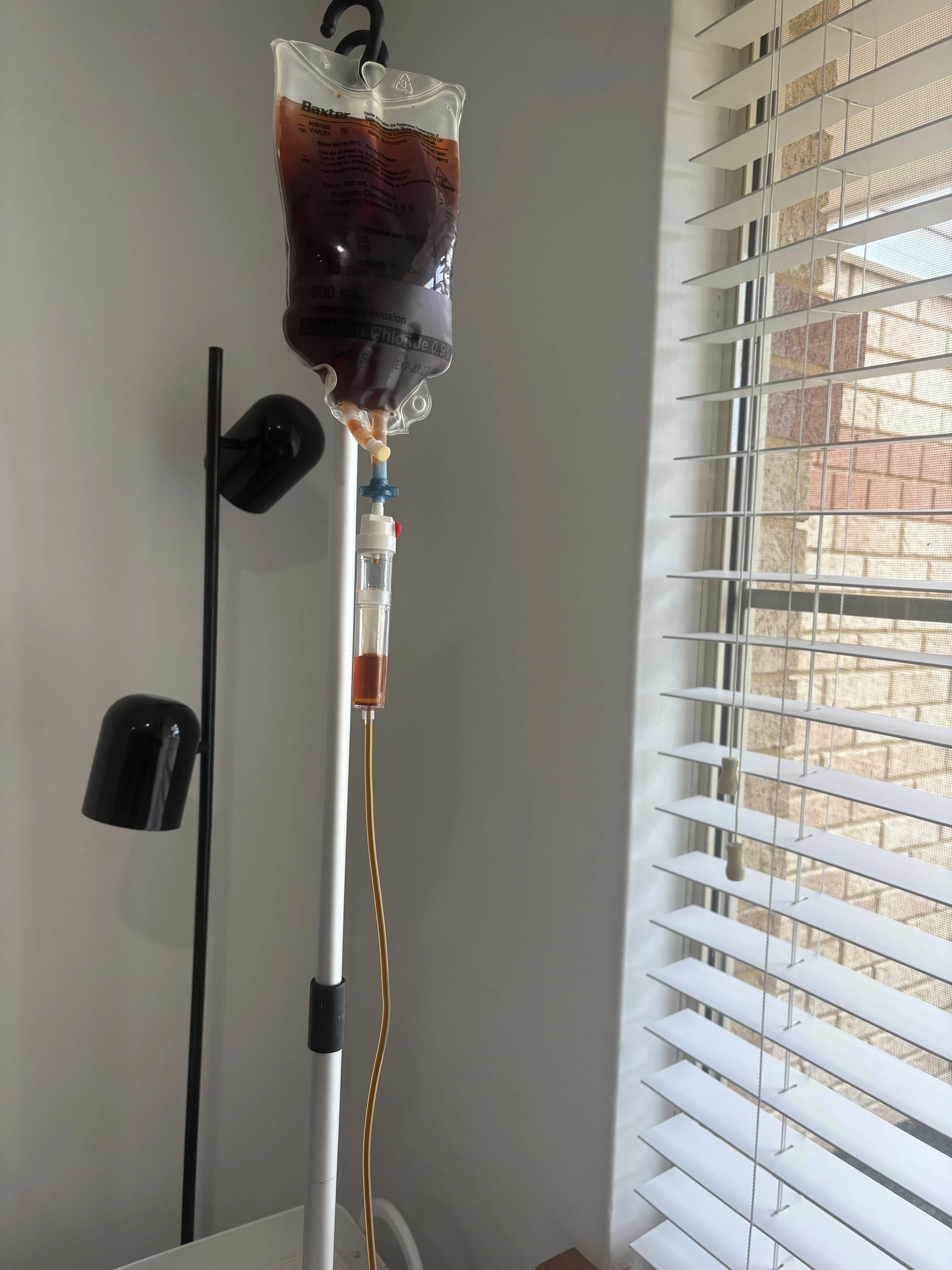 A medical IV drip bag filled with blood connected to a stand and a tube, positioned indoors near a window with open blinds, letting in natural light.