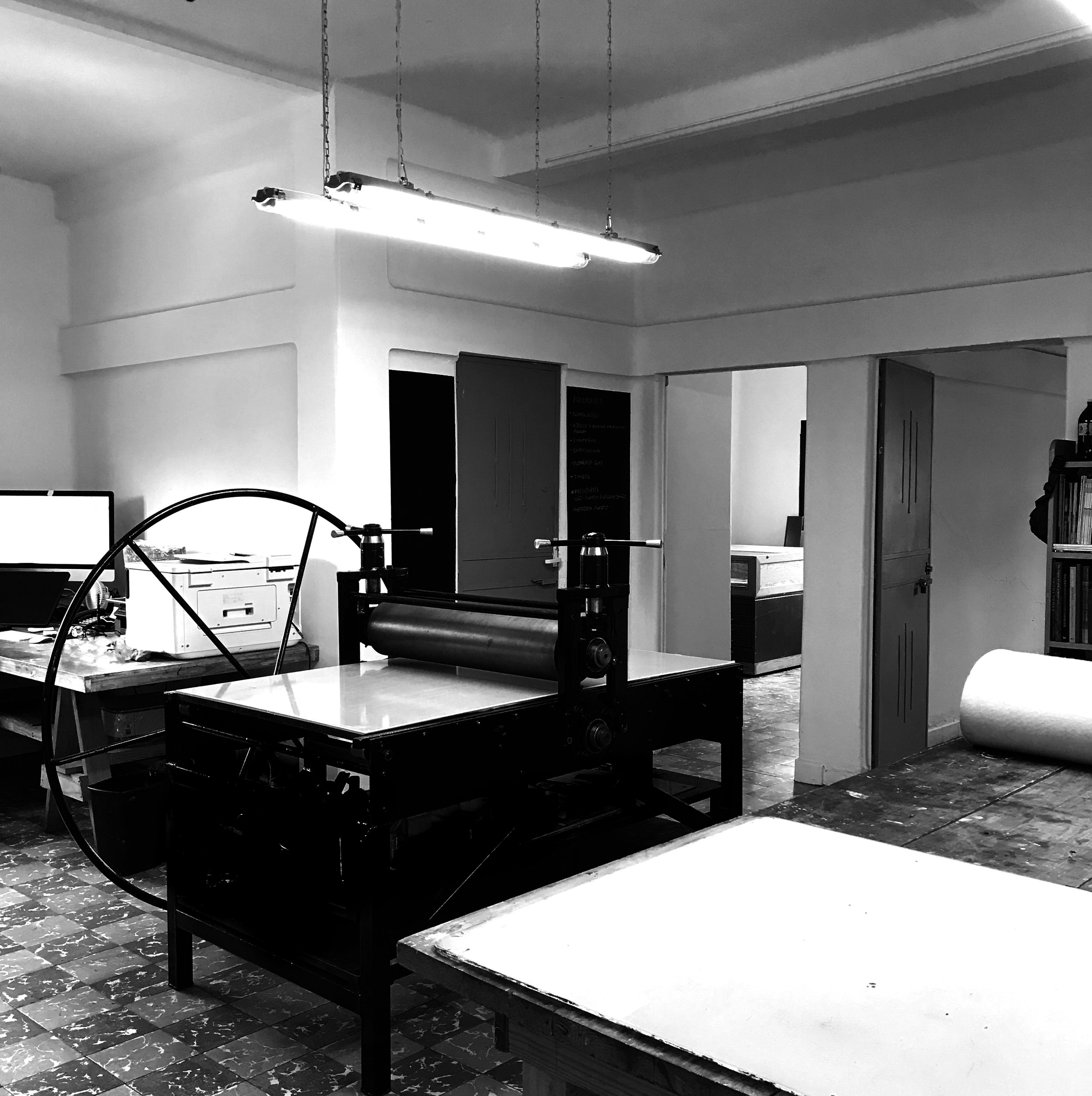 Salón interior con mesa de trabajo, equipo de impresión, y lámara suspendida en el techo. Pared blanca y puerta en el fondo.