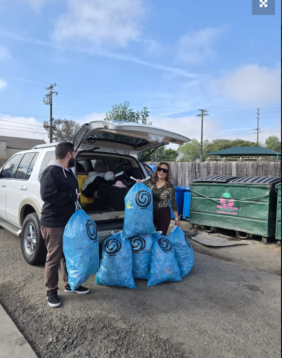 Two people, a man and a woman, unloading blue shopping bags from an open SUV in a parking lot. The woman is wearing sunglasses and a leopard print top, while the man is dressed in a black hoodie and brown pants. There is a large green dumpster and a wooden fence in the background, along with a partly cloudy sky.