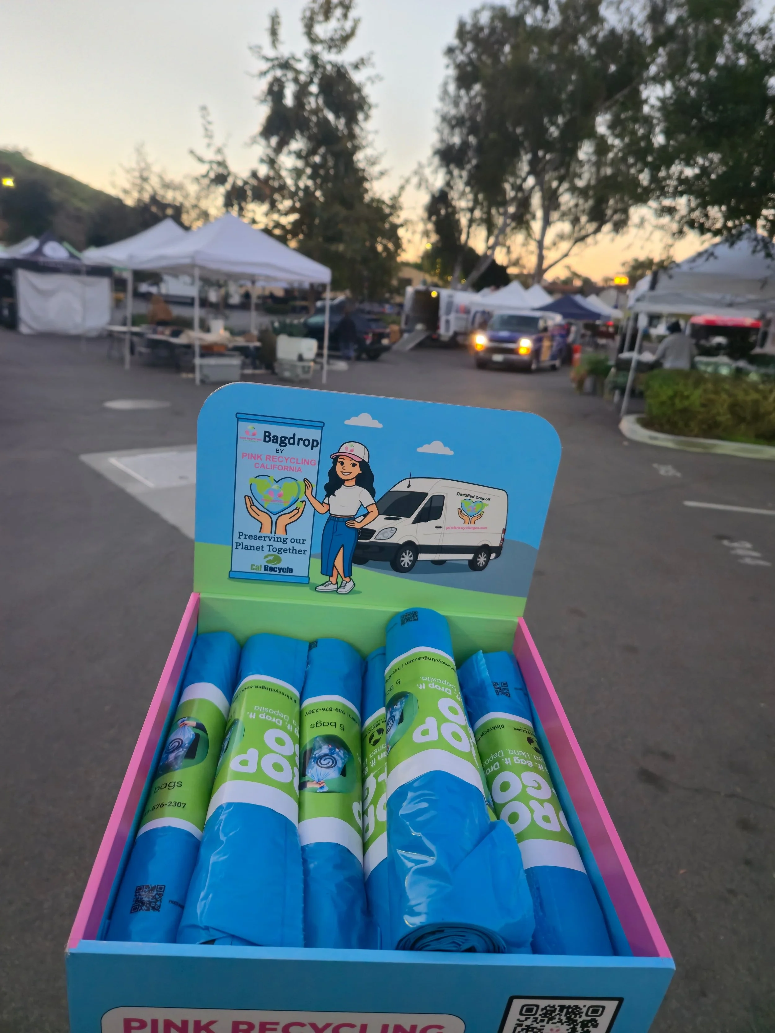 A pink and blue recycling bin with tubes of biodegradable trash bags, and a cardboard sign promoting environmental preservation with a cartoon woman holding a recycling bin, set outdoors during sunset with tents and trees in the background.