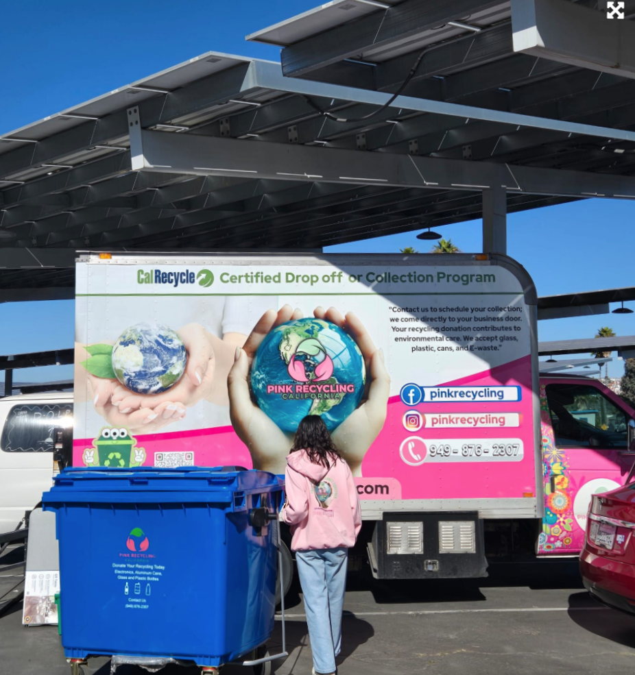 A woman drops off recycling at a pink recycling truck under a canopy in a parking lot. The truck promotes a certified drop-off or collection program and features images of the Earth being held in hands, along with social media contact information.