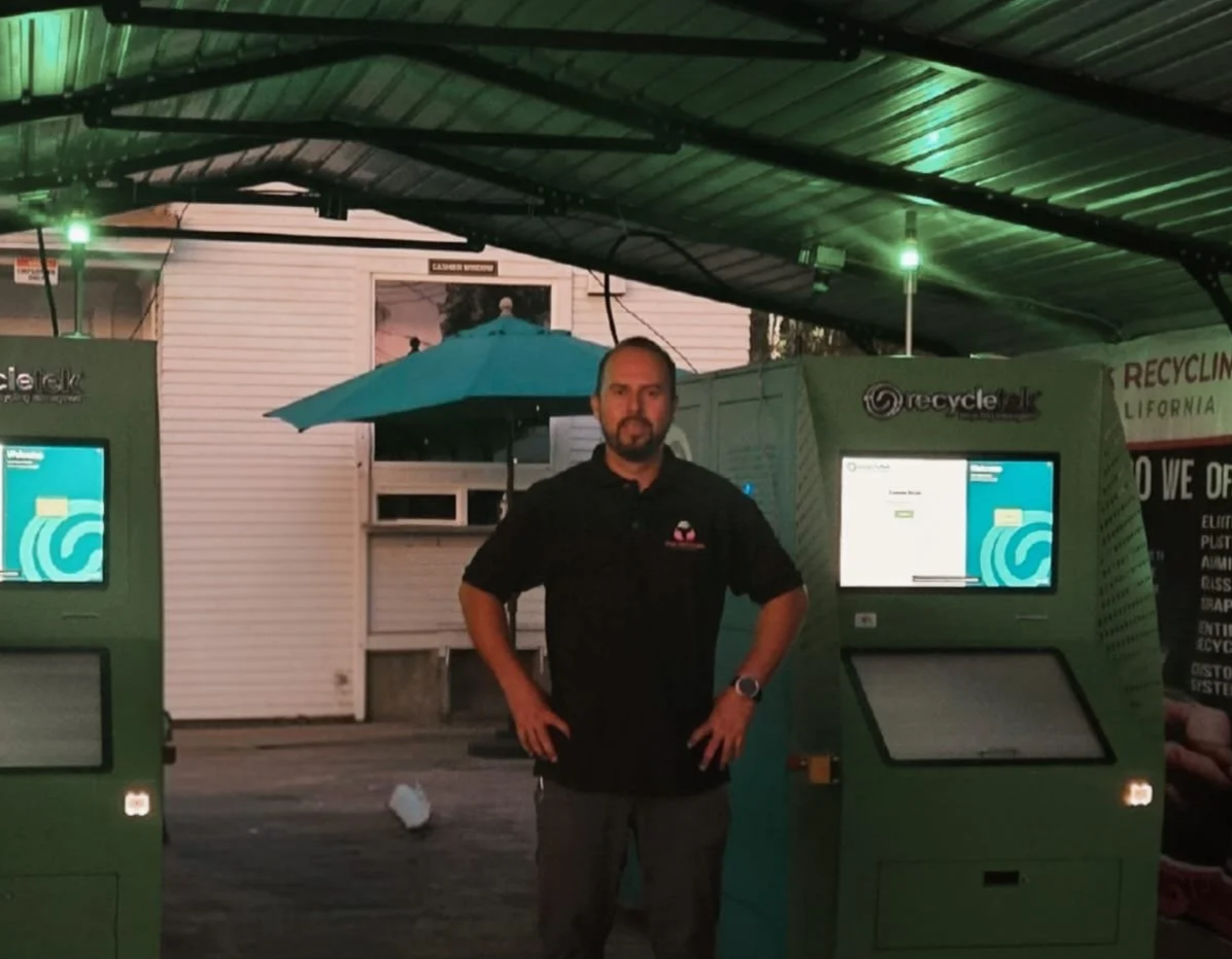 Man standing between two recycling kiosks under a roof, with a large outdoor umbrella behind him, in front of a white building with a window.