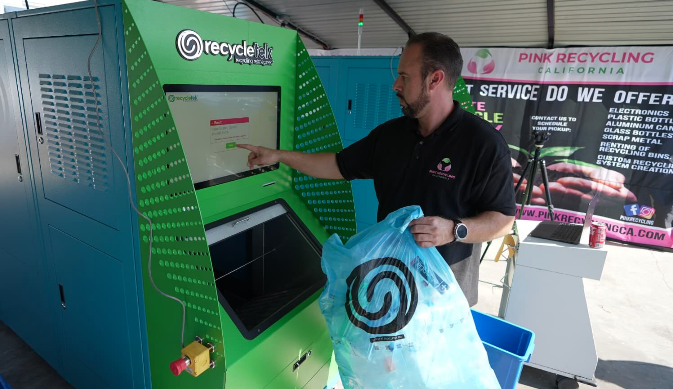A man with a beard and wearing a black polo shirt is using a recycling vending machine to recycle a plastic Target shopping bag. The machine has a large digital screen displaying an error message. In the background, there's a banner for Pink Recycling California and a small table with a laptop, a can of soda, and a bicycle.