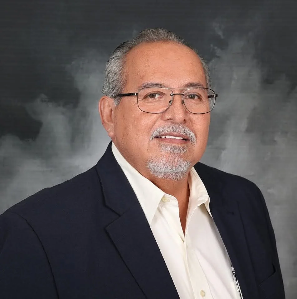 A middle-aged man with gray hair and beard, wearing glasses, a white shirt, and a dark blazer, posing against a dark, textured background.