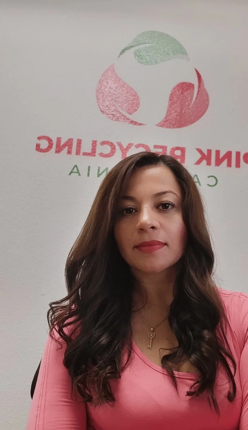 A woman with long brown wavy hair wearing a pink top and a gold necklace taking a selfie in front of a wall with a logo and text.
