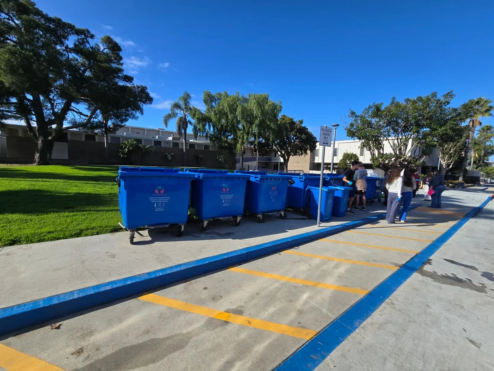 Blue recycling bins lined up on a parking lot sidewalk, with a group of people gathered nearby and trees and buildings in the background.