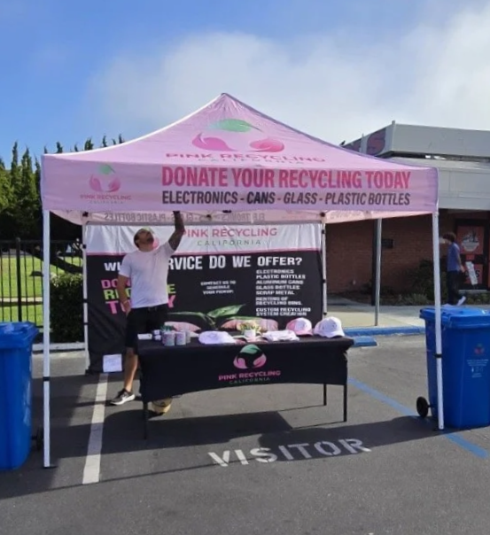 A person standing behind a recycling booth under a pink canopy with signs promoting recycling of electronics, cans, glass, and plastic bottles in a parking lot.