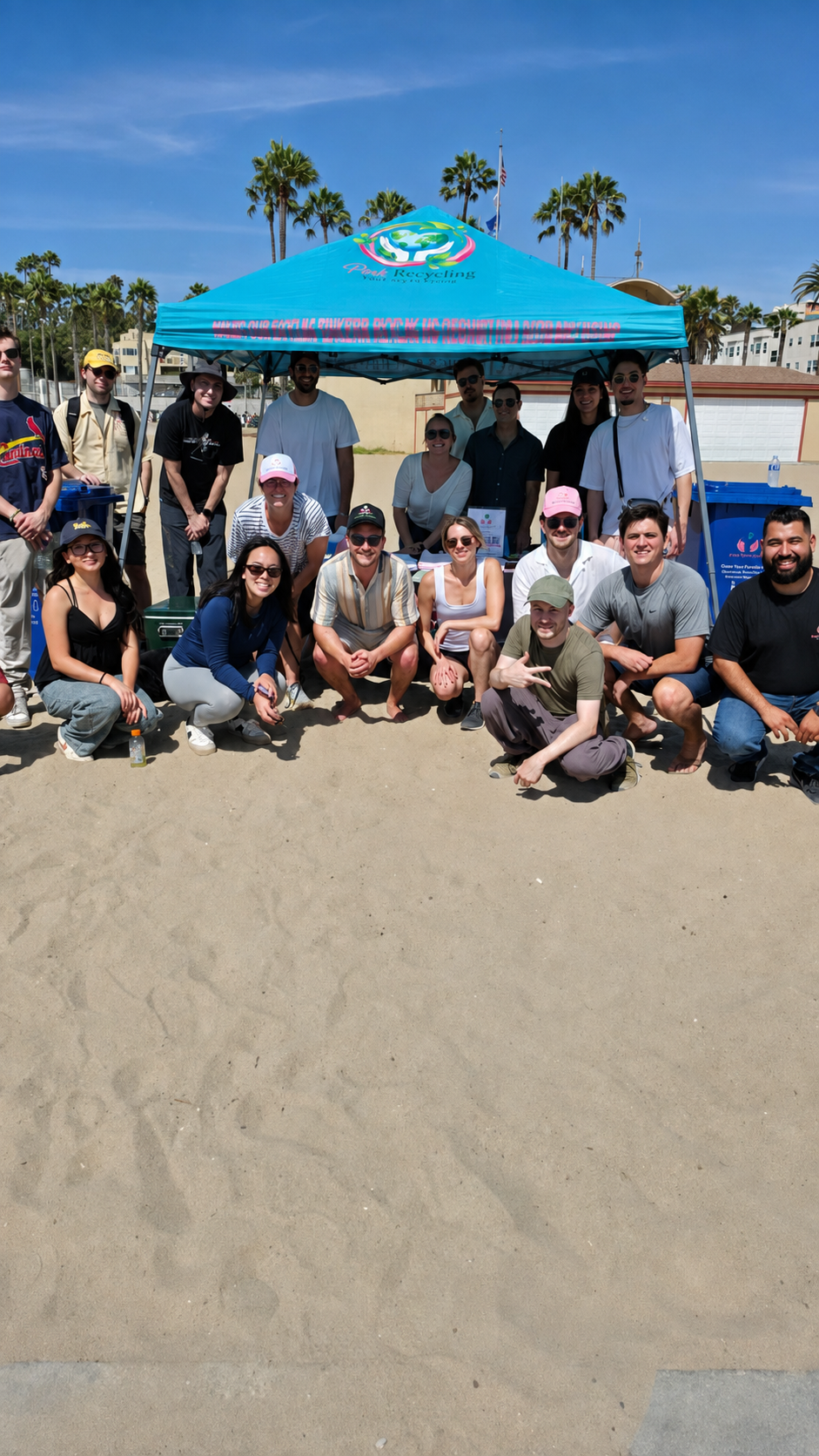 Group of people gathered under a blue canopy tent on a sandy beach, with palm trees and buildings in the background, participating in an outdoor environmental cleanup event.