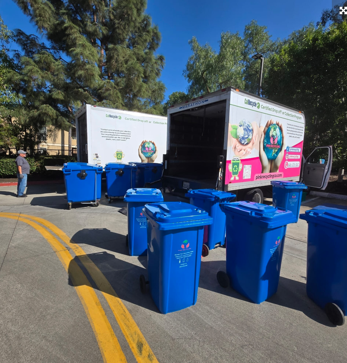 A mobile recycling truck and several blue recycling bins outside on a sunny day, with trees and a building in the background.