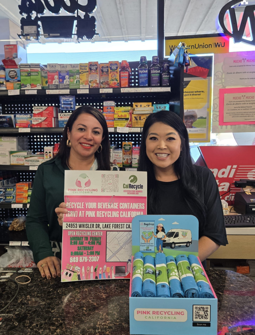 Two women at a store counter holding recycling information posters, with a display of rolled reusable bags labeled PINK RECYCLING CALIFORNIA in front of them.
