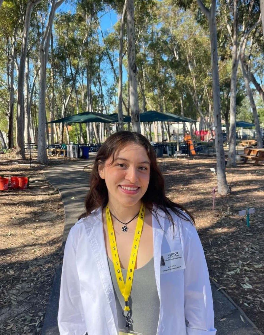 A young woman smiling outdoors in a wooded area, wearing a white shirt, a visitor badge, and a yellow visitor lanyard.