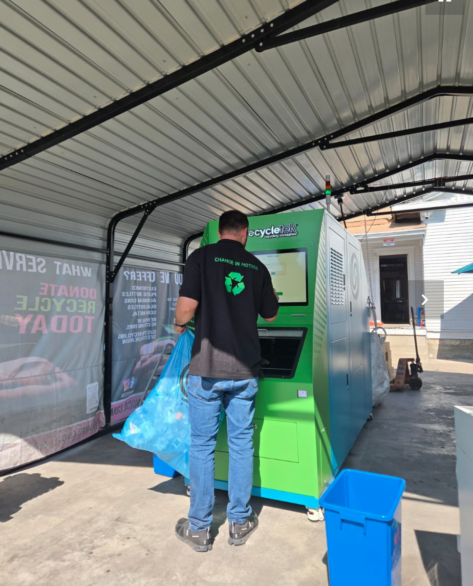 A man using a green RecyclingTek vending machine under a metal carport. The man is holding a blue plastic bag, likely for recycling. There is a blue trash bin in front of him, and the vending machine has a touchscreen interface. The background shows a residential house and some equipment.