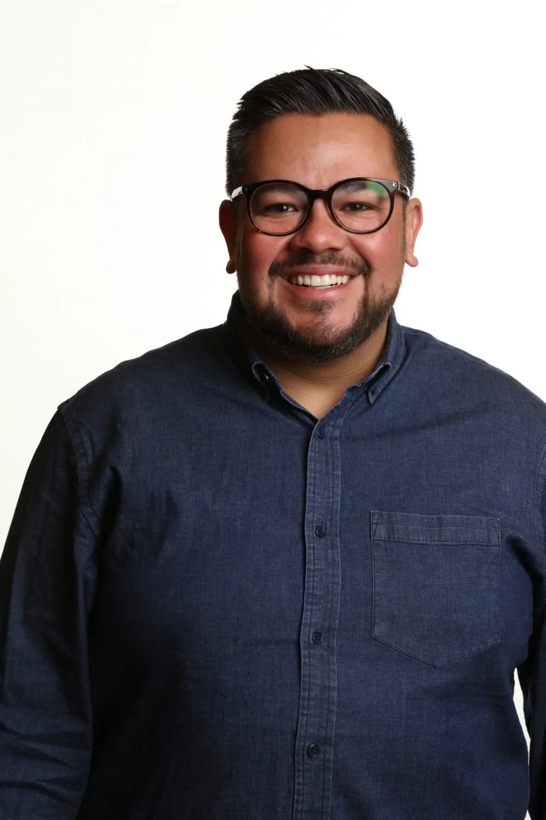 Portrait of a smiling man with glasses, short dark hair, beard, wearing a dark blue button-up shirt, against a plain white background.