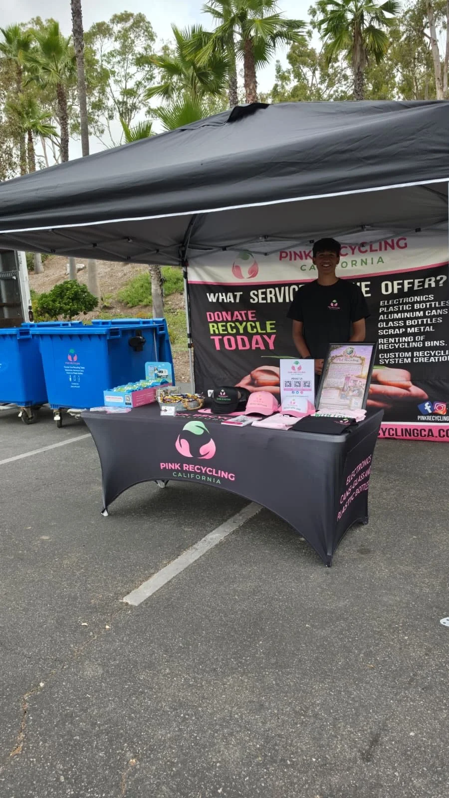 A booth at an outdoor event promoting Pink Recycling California, featuring a table with informational materials and a person standing behind it, under a canopy with trees in the background.