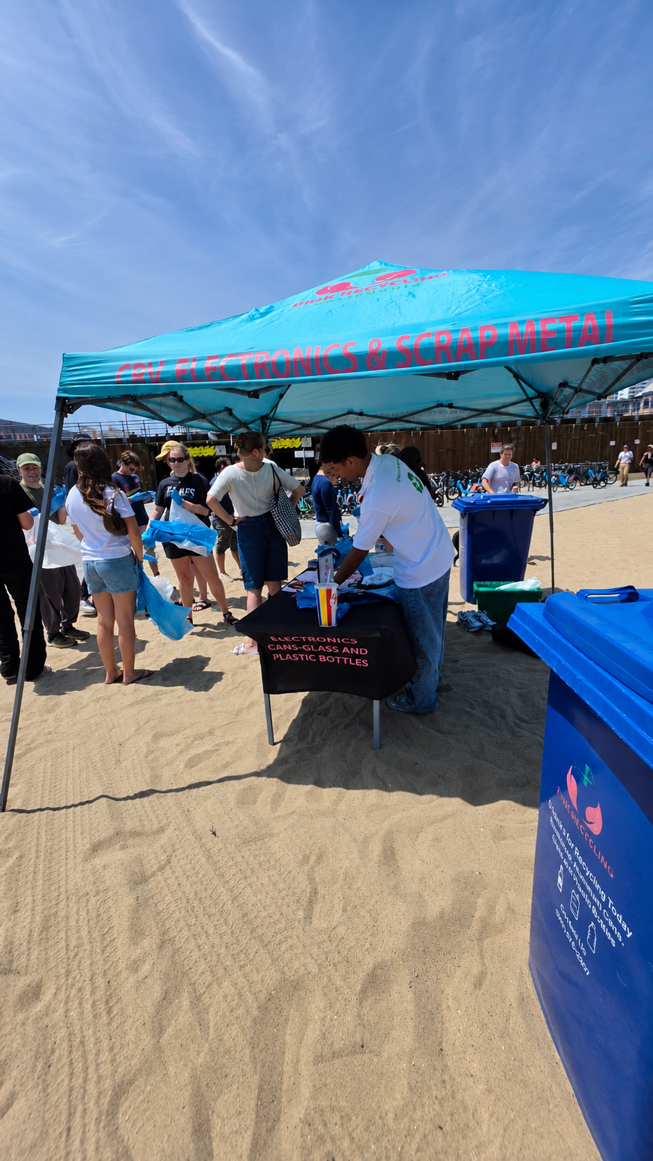 People gathered under a blue tent labeled "CRV Electronics & Scrap Metal" at an outdoor event on a sandy area, with waste disposal bins nearby. The tent's table displays electronics, cans, glass, and plastic bottles, and an individual is assisting visitors.