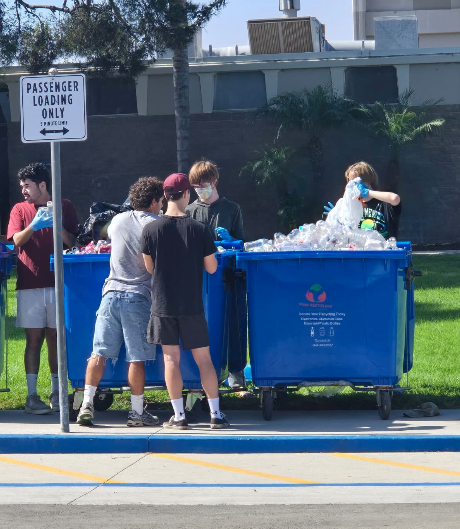 Group of five young people recycling plastic bottles into large blue recycling bins outdoors near a sidewalk, with a street and building in the background.