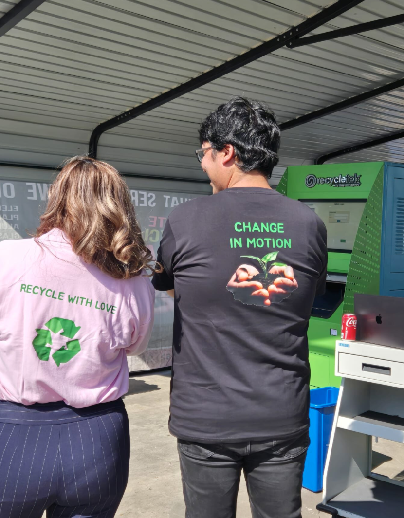 Two people at a recycling station, one wearing a pink shirt with the green recycling symbol and the phrase 'Recycle with Love', and the other wearing a black shirt with the phrase 'Change in Motion' and an image of a hand holding a small plant. The recycling station is green with a logo and the word 'recycletek' on it.
