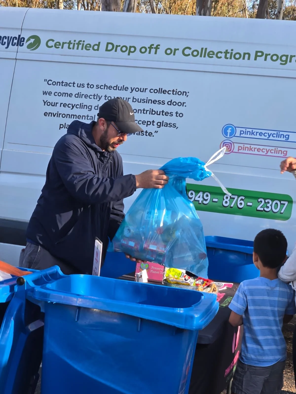 Man wearing a black cap and sunglasses holding a large blue recycling bag filled with cans and bottles at a recycling event. There is a large white vehicle in the background with green and black text about recycling drop-off programs and social media links. A young boy in a striped shirt is standing near a table with snacks and other items.