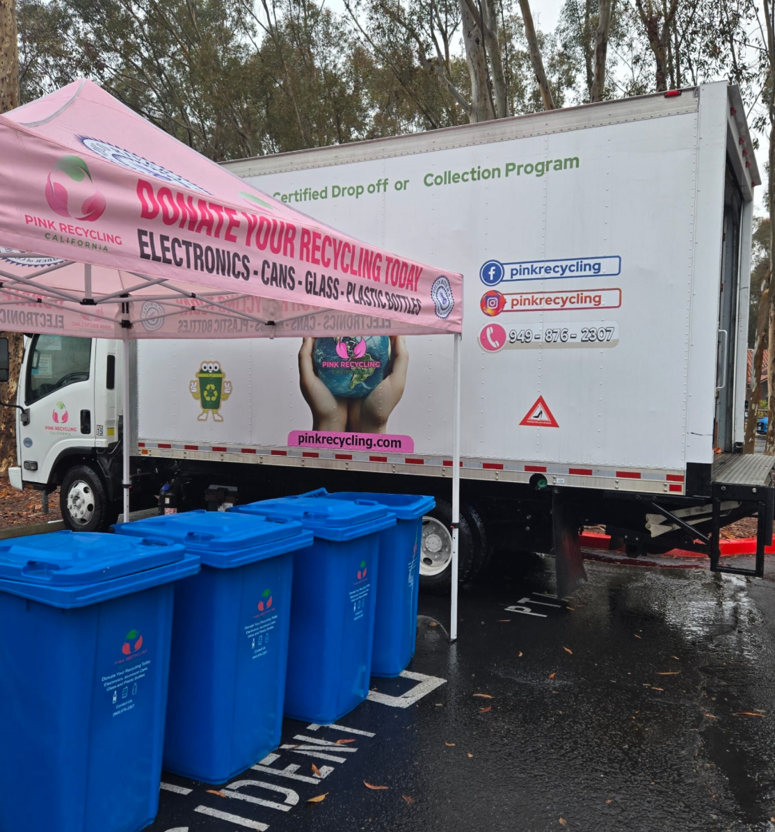 A mobile recycling truck with social media and contact info, a pink recycling tent, and four blue recycling bins on a wet parking lot.