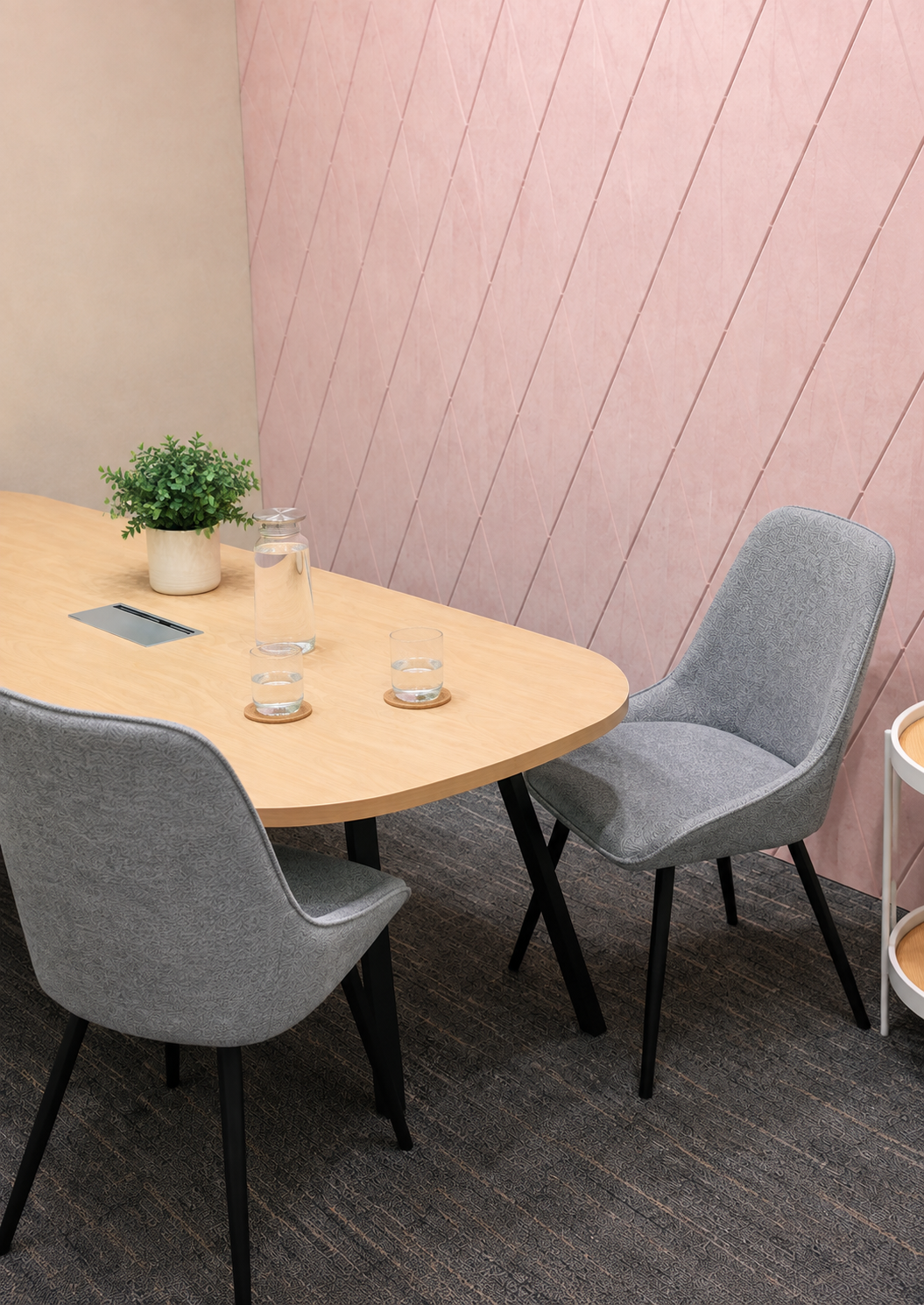 A conference room with a light wooden table, three grey chairs, a small plant, a glass water pitcher with two glasses, and a pink textured wall.