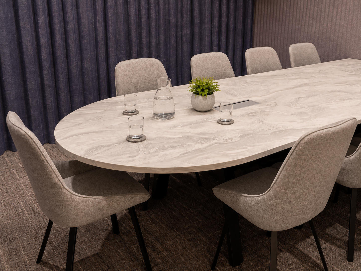A modern conference room with a large oval marble table, surrounded by six gray upholstered chairs. On the table are a glass pitcher of water, four glasses, a small potted green plant. The background features dark purple curtains and a beige textured wall.