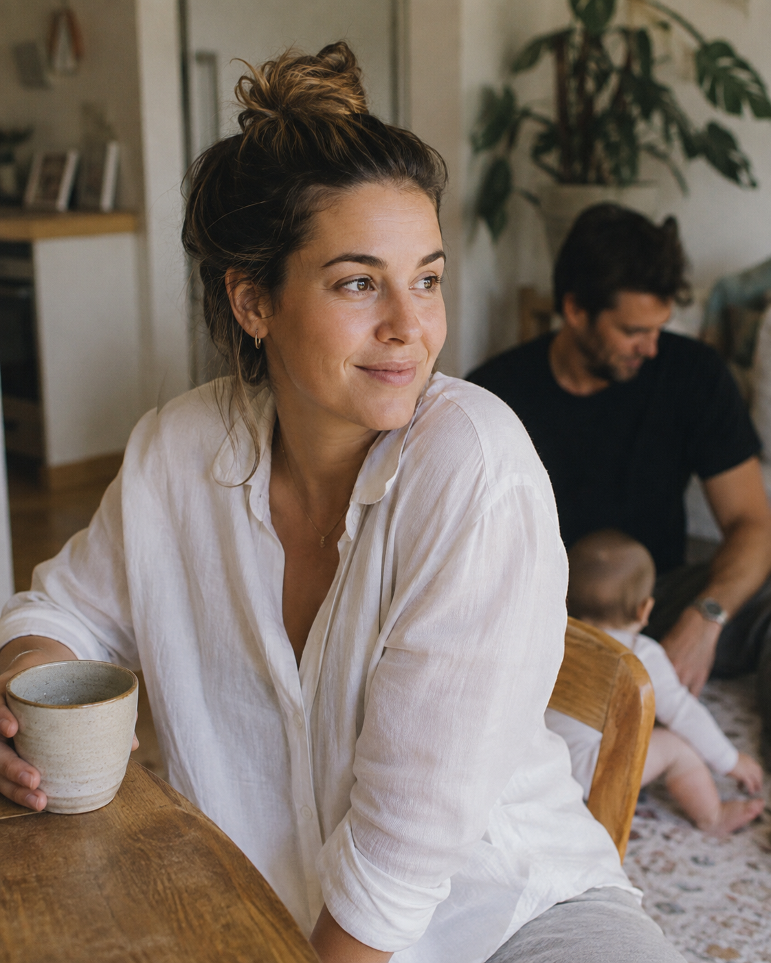 A woman with brown hair tied in a bun, wearing a white shirt, sitting at a wooden table holding a mug, with a man and a child in the background in a cozy, well-lit room.