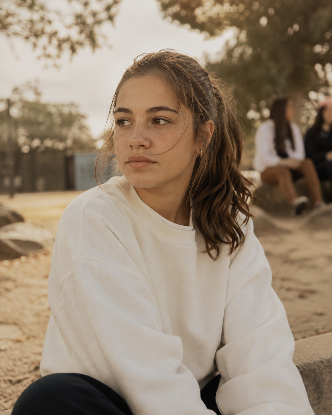 A young woman with brown hair in a ponytail, wearing a white sweatshirt, sitting outdoors near trees with two women in the background on a bench.