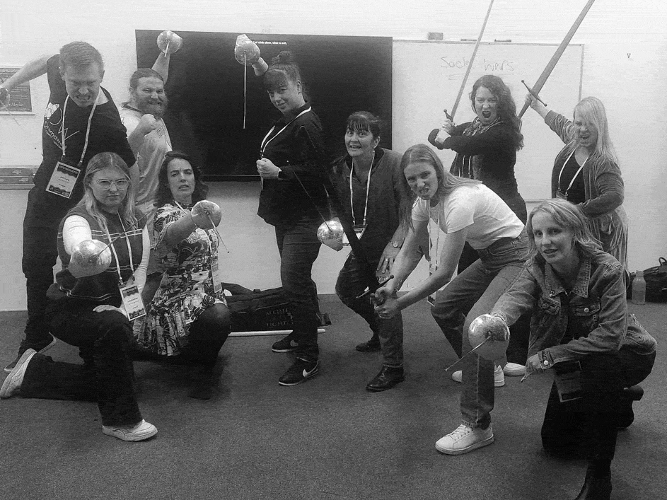 Group of people posing with swords in a stage fighting workshop, some kneeling or crouching.