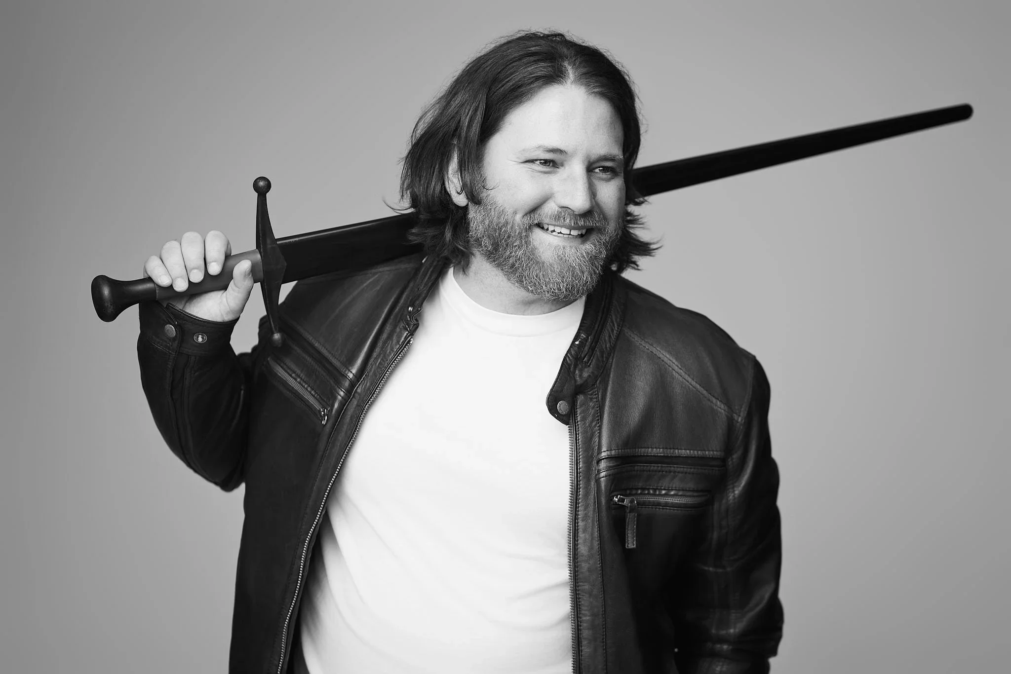 A man smiling while holding a sword over his shoulder, wearing a leather jacket and white shirt, against a plain background.