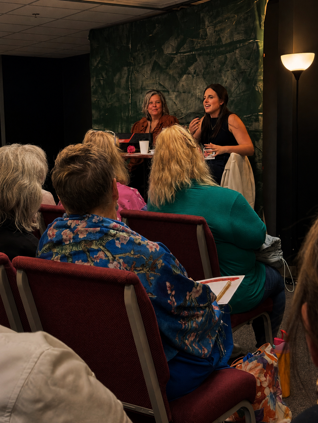 Two women are speaking on a stage with an audience of mostly women seated in red chairs. One woman is wearing a black sleeveless top, and the other is wearing a brown jacket. The setting appears to be an indoor event or seminar with dark green backdrops and soft lighting.