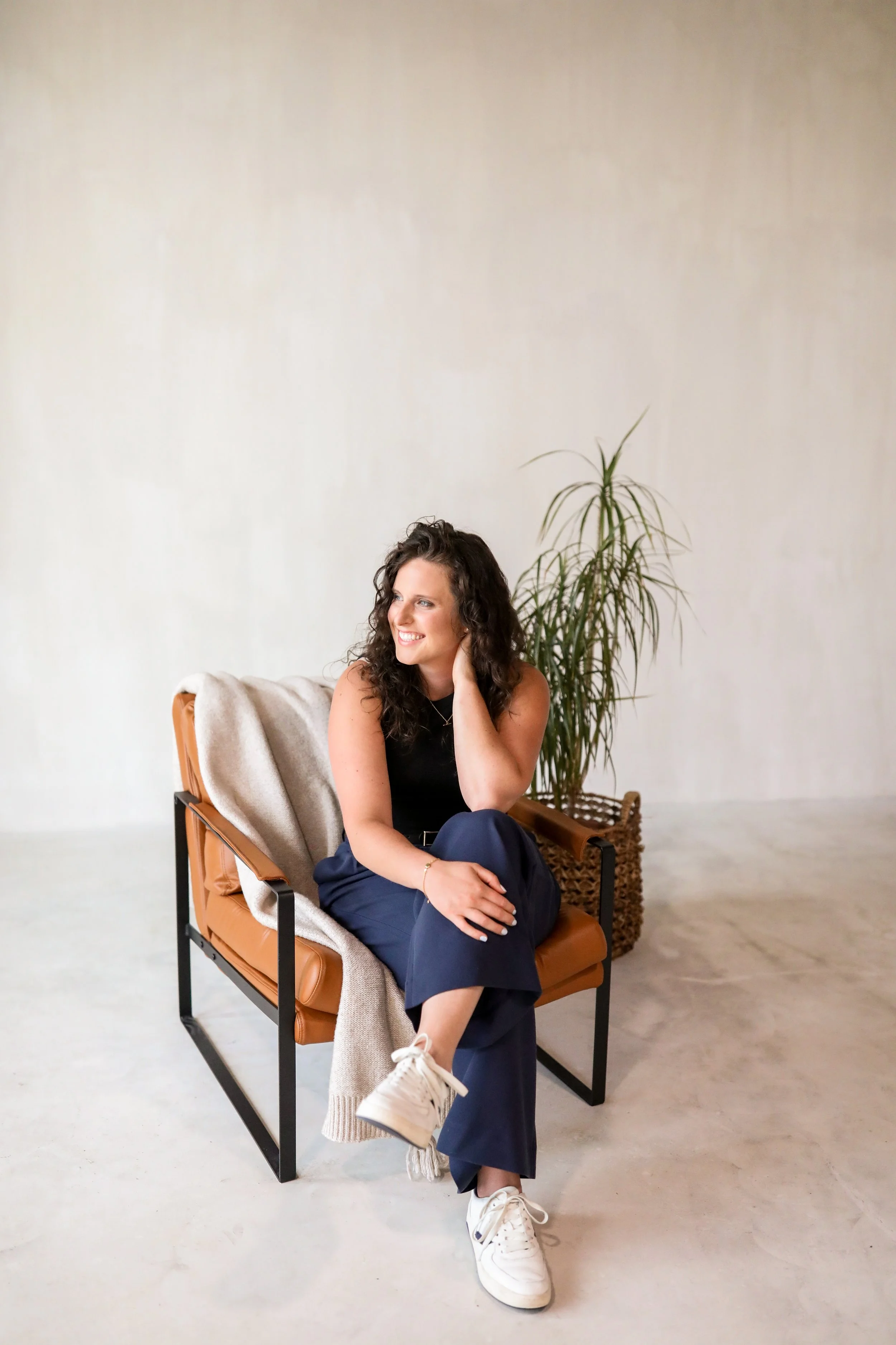 A smiling woman with curly brown hair sitting on a modern tan and black chair with a blanket on the armrest, in a minimalist room with a large green potted plant behind her, and a plain light-colored wall.