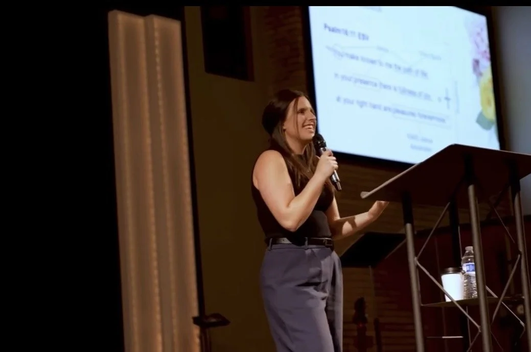 A woman standing at a podium speaking into a microphone in front of a large presentation screen.