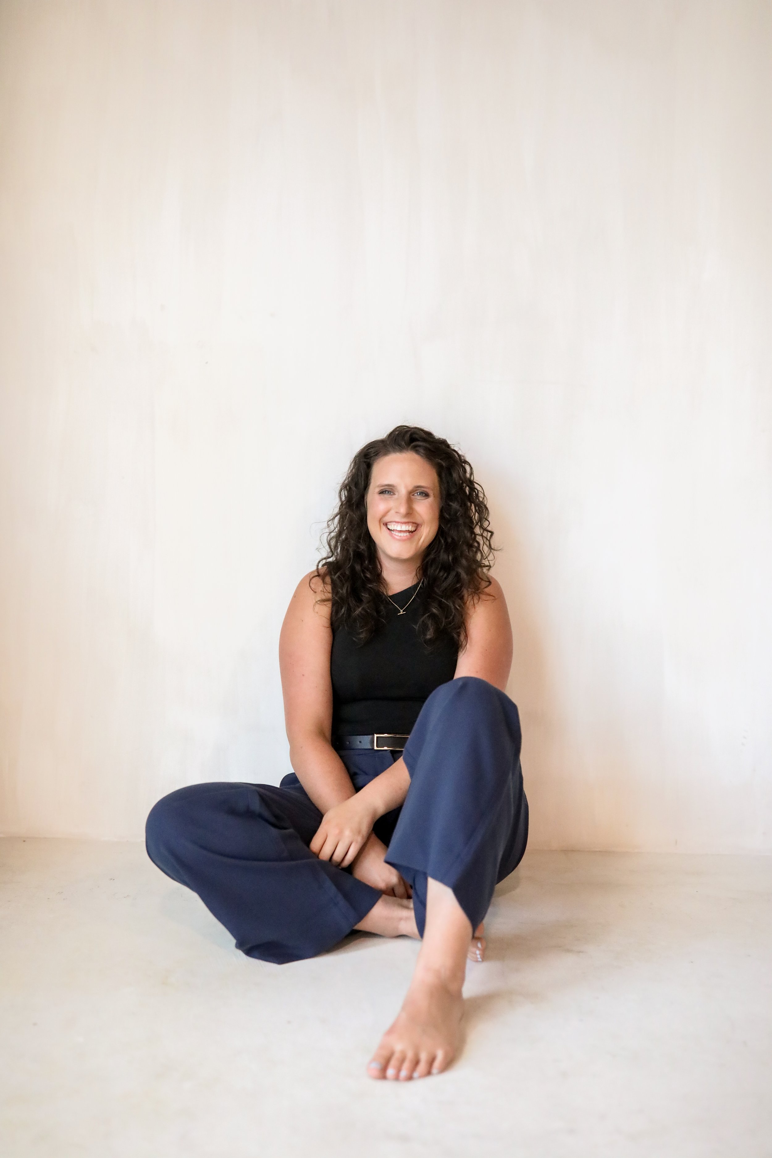 A woman with curly dark hair sitting on a light-colored floor against a plain off-white wall, smiling, wearing a black sleeveless top and navy blue pants, with one leg crossed and her foot visible.