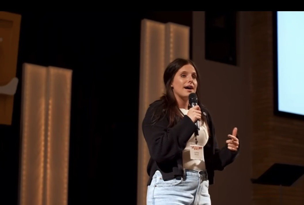 A woman holding a microphone speaking on stage at a conference or event.