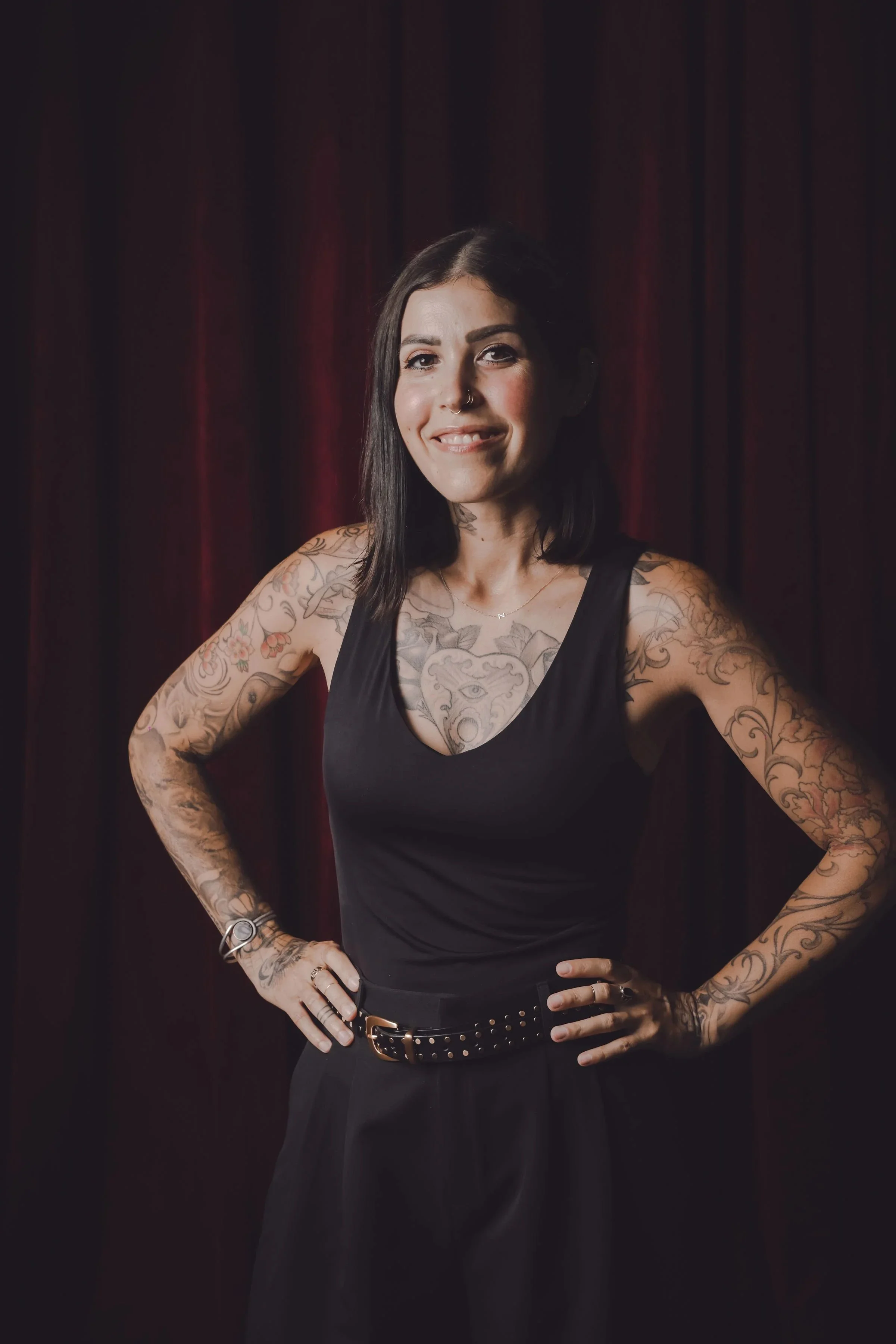 A woman with shoulder-length black hair, tattoos on her arms and chest, wearing a black tank top and black pants, standing with her hands on her hips in front of a dark red curtain, smiling slightly with one eye closed.