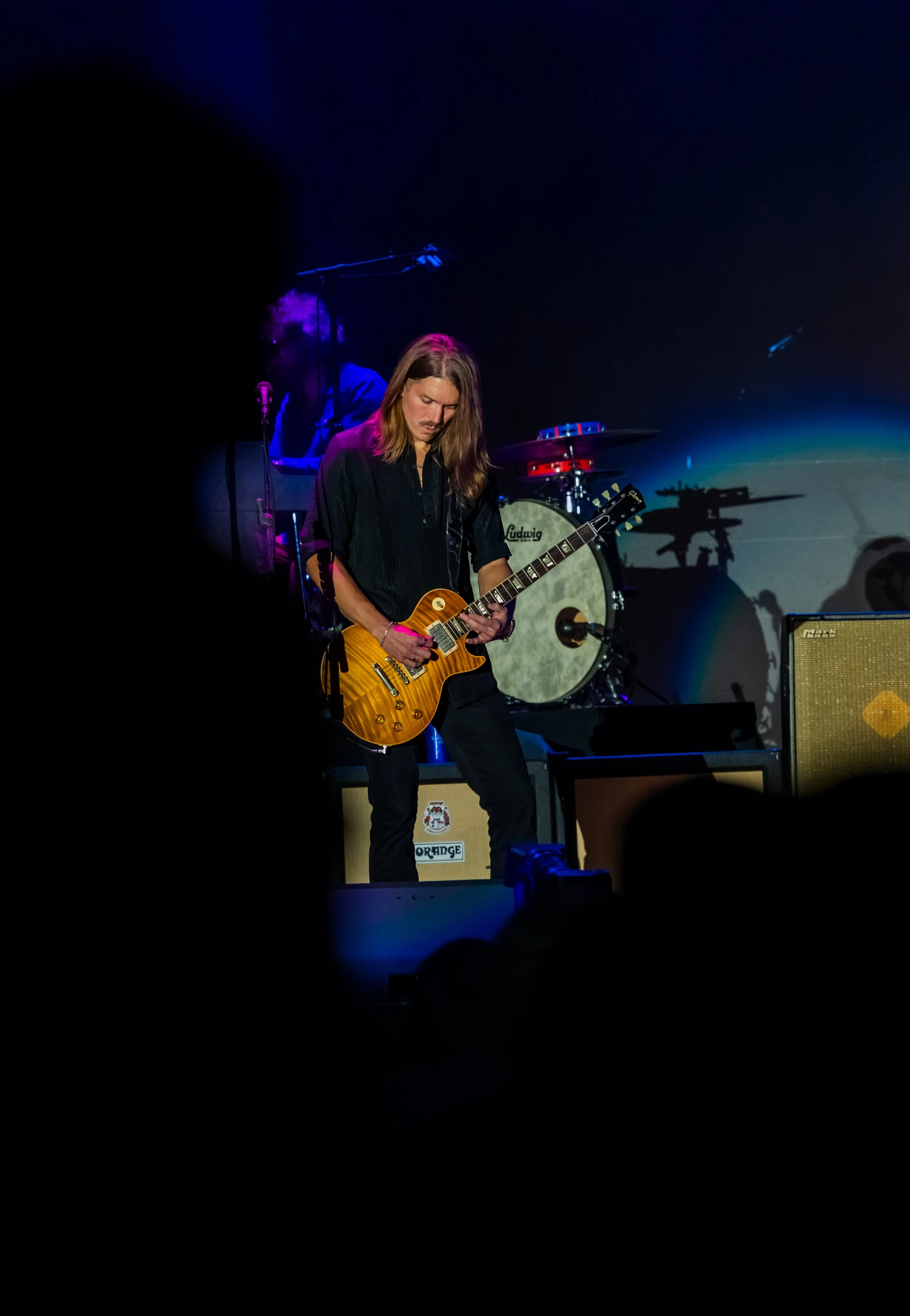 A male guitarist with long hair playing an electric guitar on stage, with a drum set and a musician in the background, illuminated by stage lights.