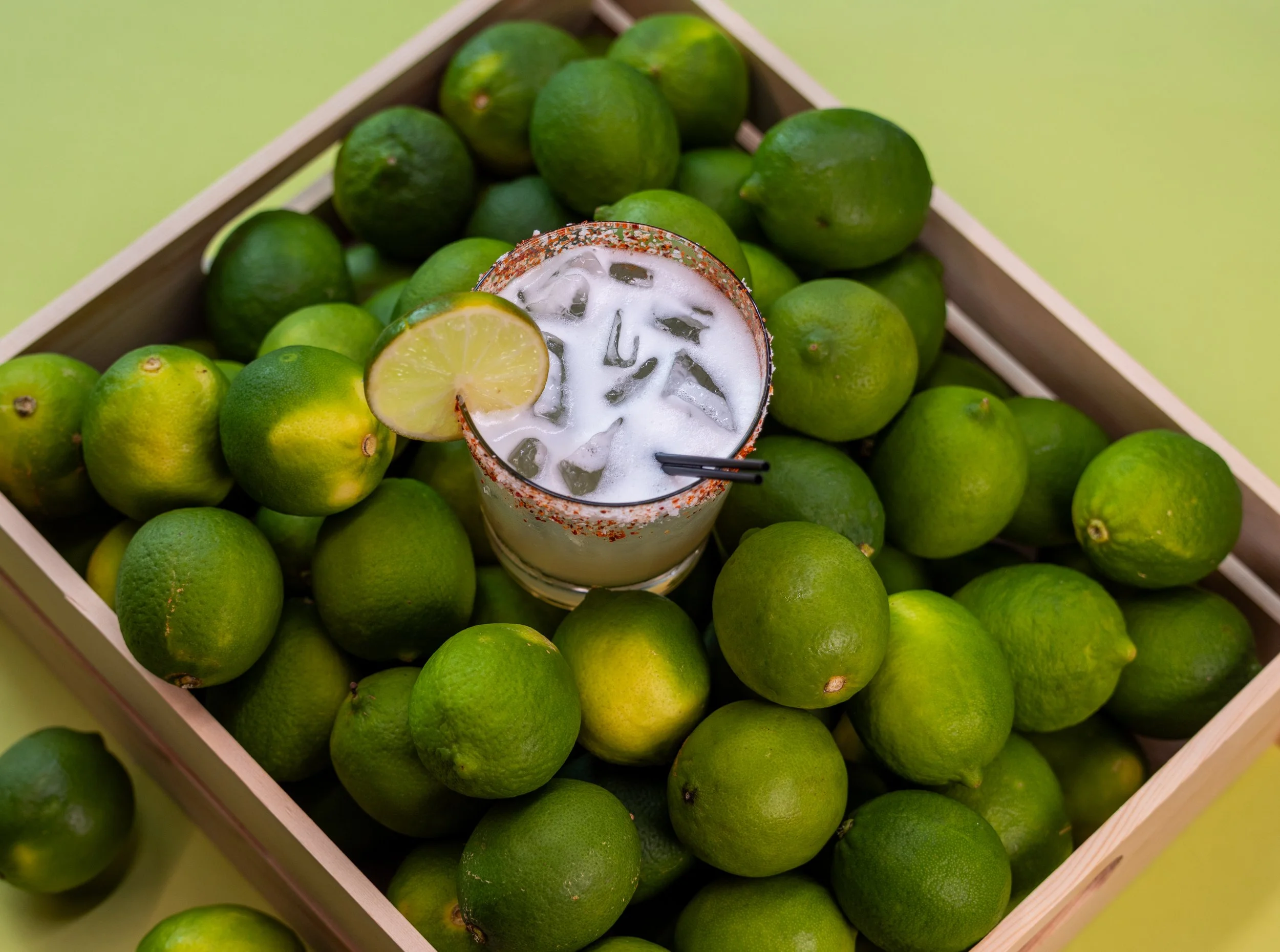 A wooden crate filled with fresh green limes, with a glass of lime-flavored drink with ice and a lime wedge on the rim placed among the limes.