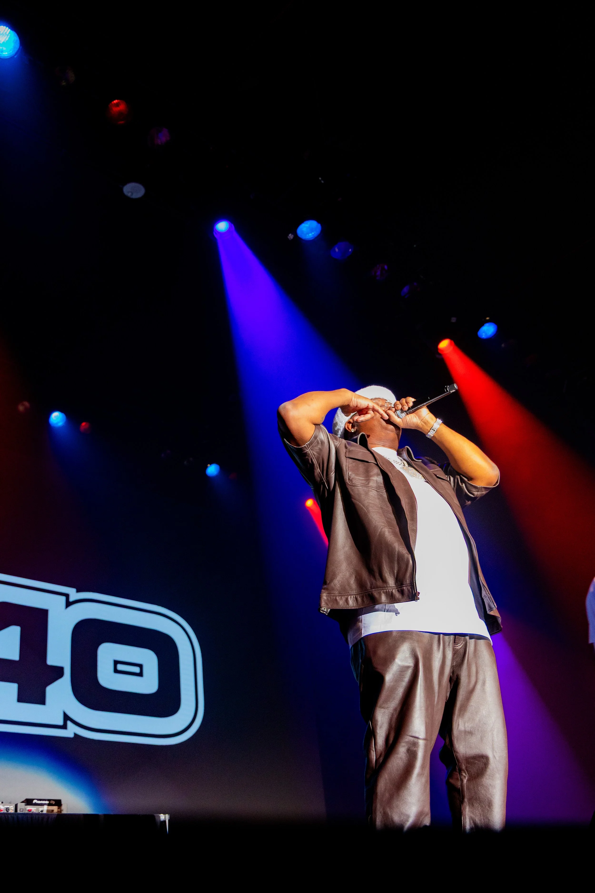 Performer singing into a microphone on stage with colorful spotlights and a logo in the background.