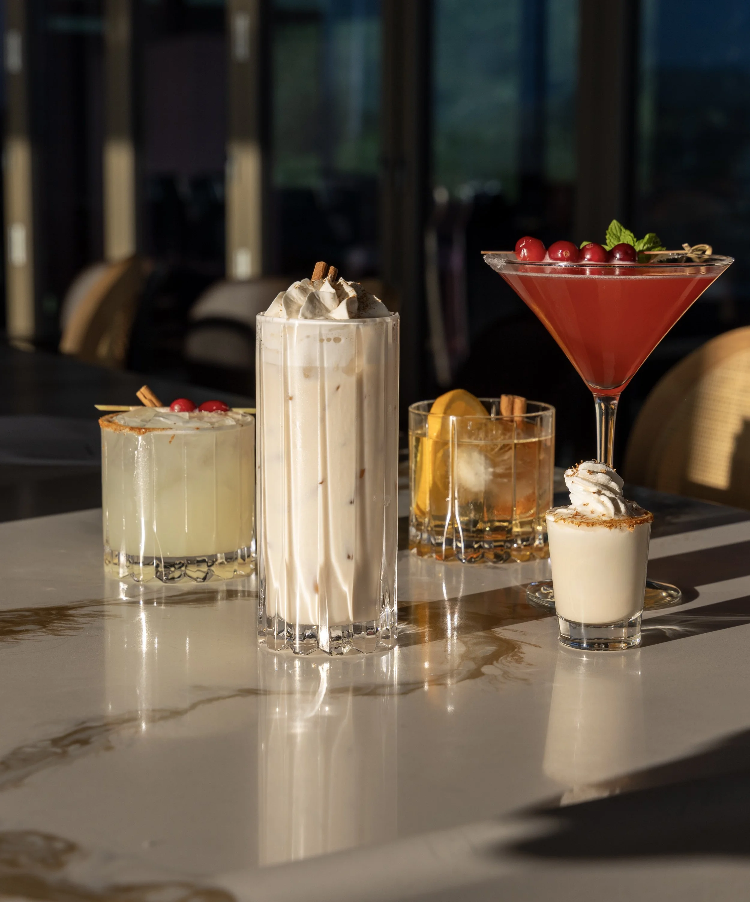 Assorted cocktails on a table with a dark background, including a milkshake topped with whipped cream and cinnamon stick, a key lime cocktail with a lime wedge, and a red fruit cocktail garnished with cherries and mint.