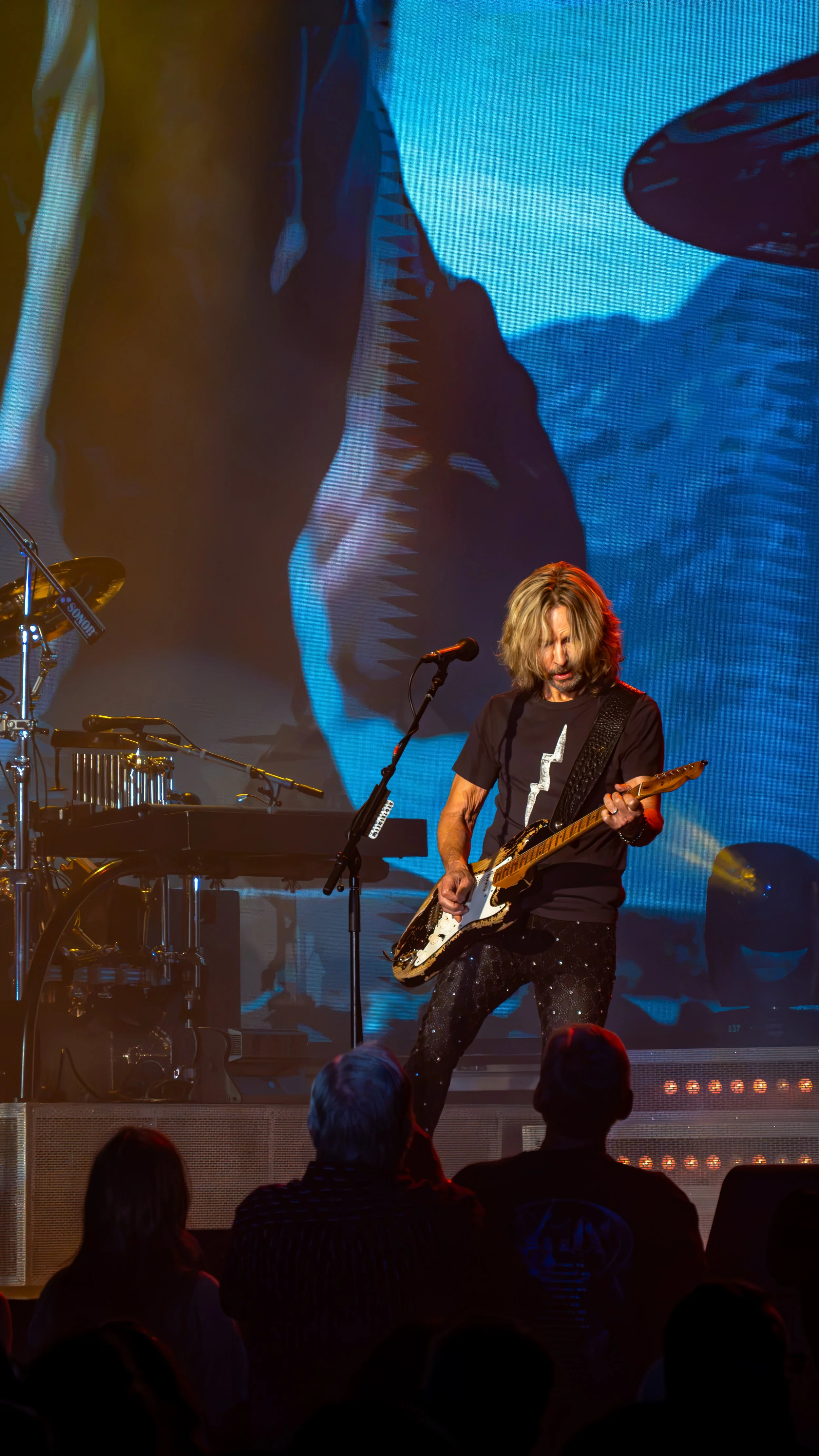 A musician with long hair playing an electric guitar on stage in front of an audience, with a large screen displaying abstract blue and black visuals in the background.
