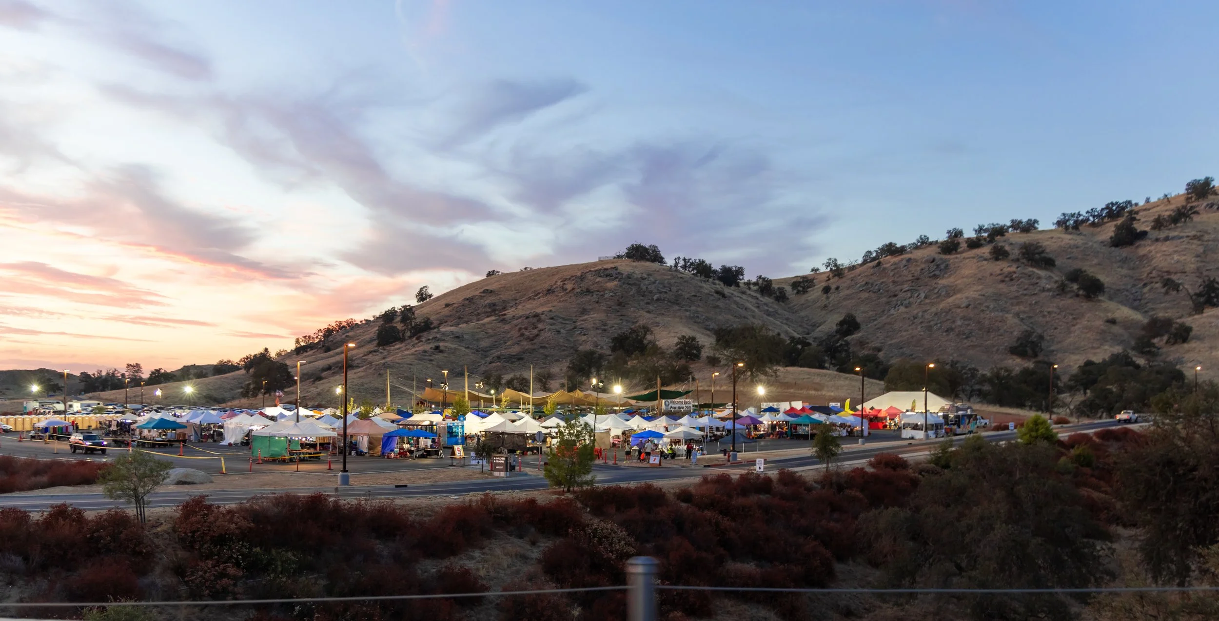 Outdoor market with tents and stalls at the base of a hillside with sparse trees, under a partly cloudy sky at sunset.