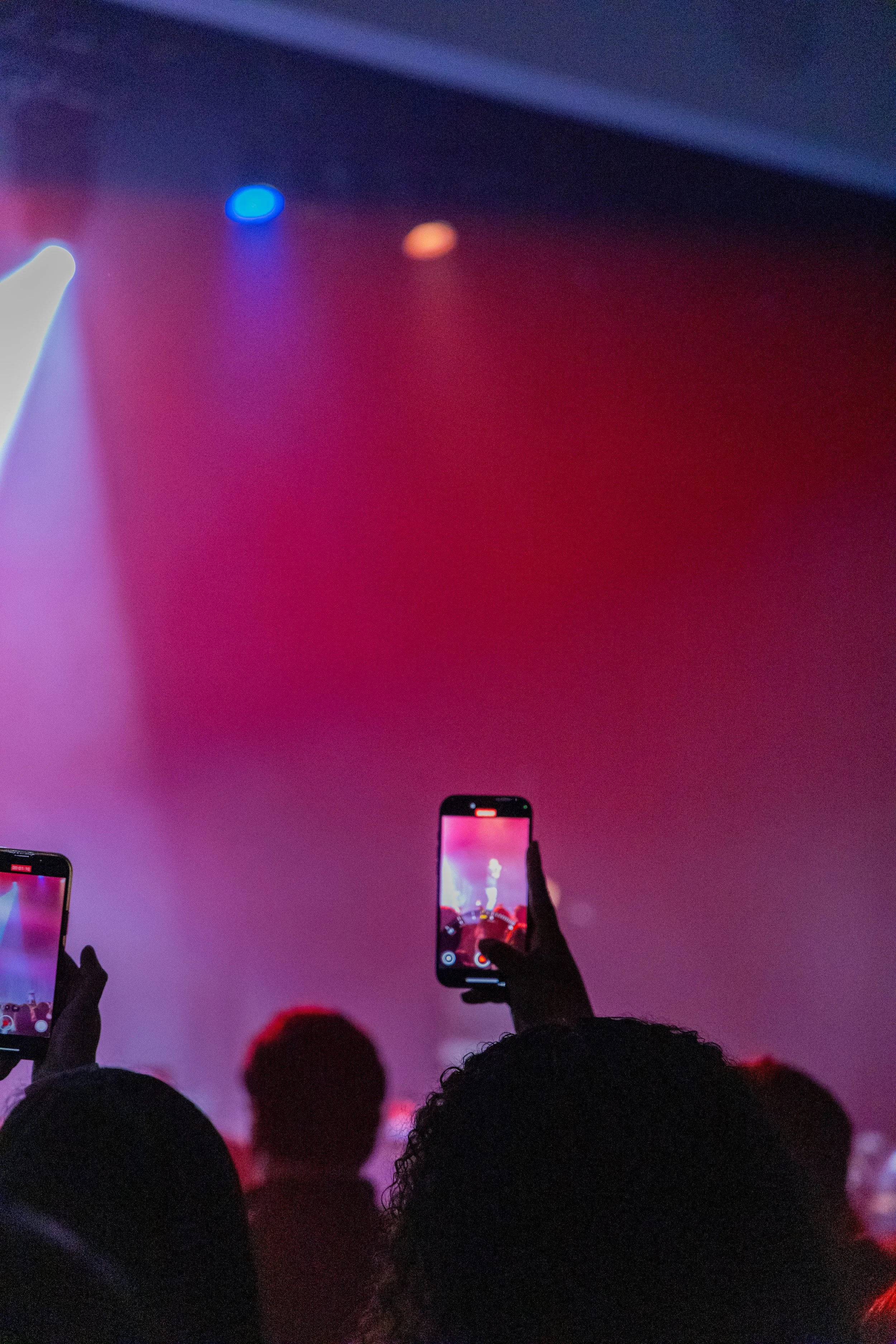People in a crowd taking photos or videos of a person performing on stage with bright, colorful lighting.