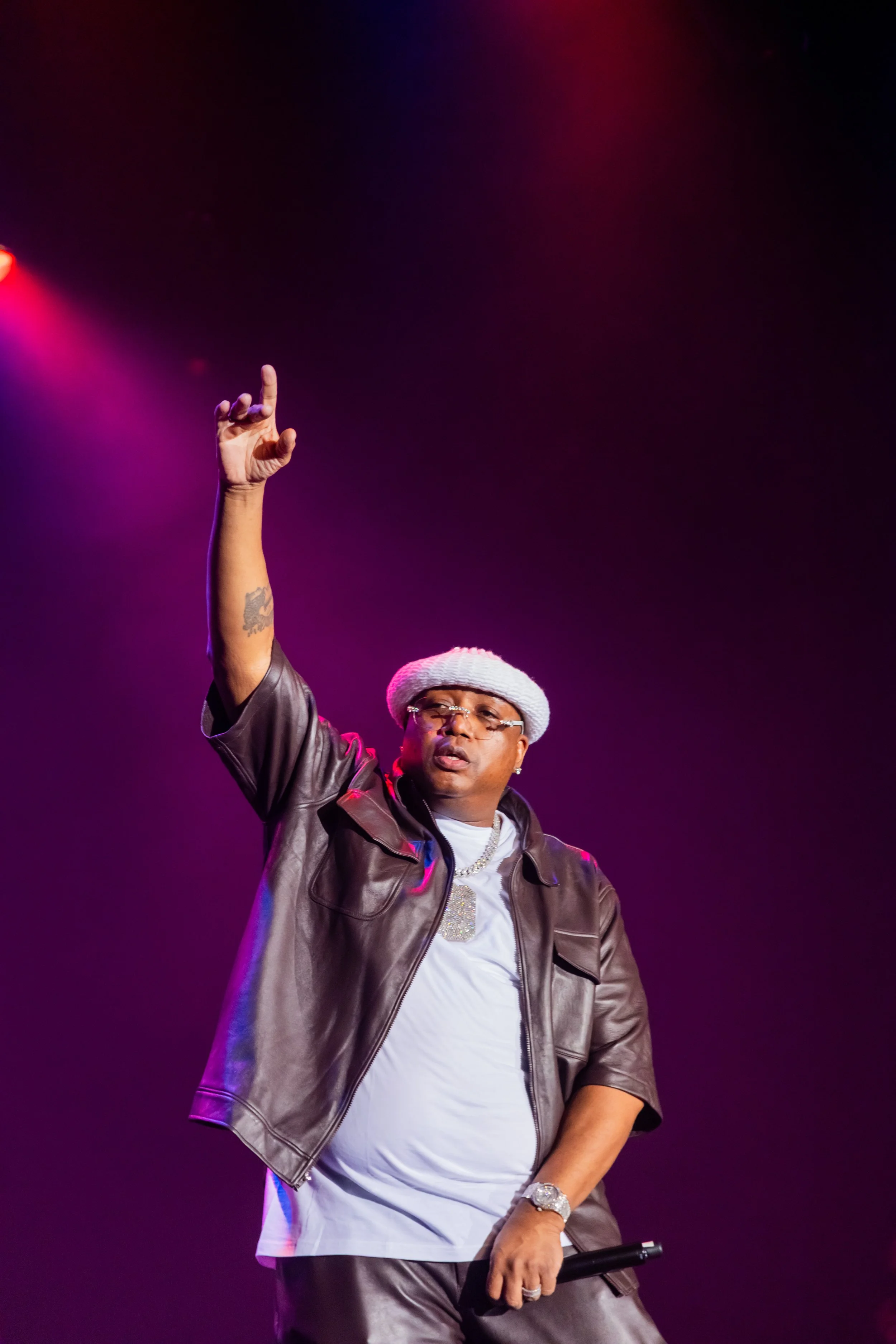 A man wearing a white beanie, glasses, a gray leather jacket, and a white T-shirt, holding a microphone, with his right arm raised, against a dark purple stage background.